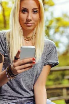 Beautiful blond girl in a park, has long hair and cell phone in hand. It could be summer or fall, but she is wearing a short-sleeved gray T-shirt.