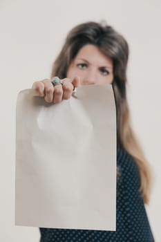 Blank sheet held by female hand with ring in foreground. The hand is of a woman with long hair and a frowning look wearing a long dark-colored dress, all this behind blurred against a white background