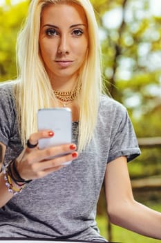 Beautiful blond girl in a park, has long hair and cell phone in hand. It could be summer or fall, but she is wearing a short-sleeved gray T-shirt.