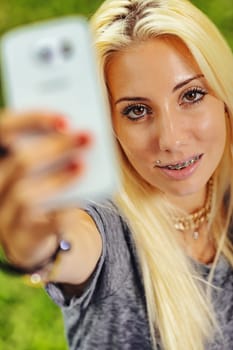 Vertical portrait of young blond woman in selfie position. Fixes the lens with intense eye-contact. She smiles highlighting her orthodontic braces that nonetheless make her even more attractive. Compl