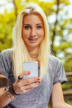 Beautiful blond girl in an outdoor forest or public park on a sunny day. She has very visible braces that make her even prettier. She has a smartphone that she has stopped looking at to turn her atten