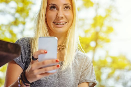 Beautiful blond girl in an outdoor forest or public park on a sunny day. She has very visible braces that make her even prettier. She is holding a smartphone, laughing and looking up beyond us.