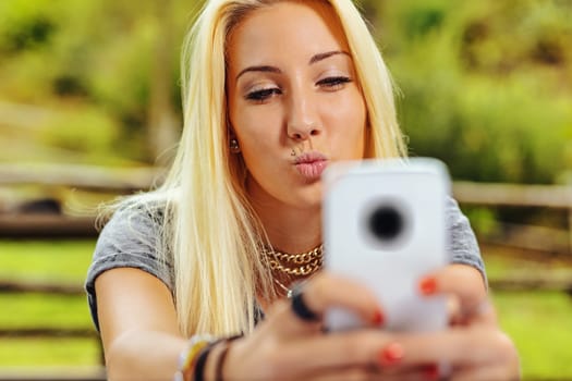 Beautiful blond girl in a park, she has long hair and holds her cell phone with two hands, toward which she sends a kiss: perhaps she is taking a selfie or is on a call with her lover