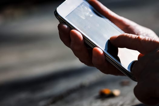 In a park, an elderly's hands rest on rustic wood, struggling with a smartphone. Tech's insensitivity to aging hands results in societal exclusion