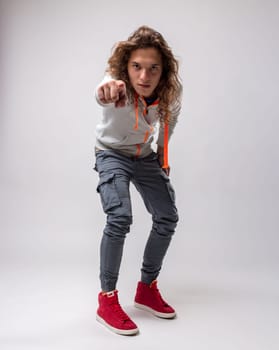 Portrait of young man with long hair. He is wearing ankle-high skinny jeans and high-top sneakers and a gray sweatshirt with neon orange details. he is standing but slightly crouched, pointing at us i