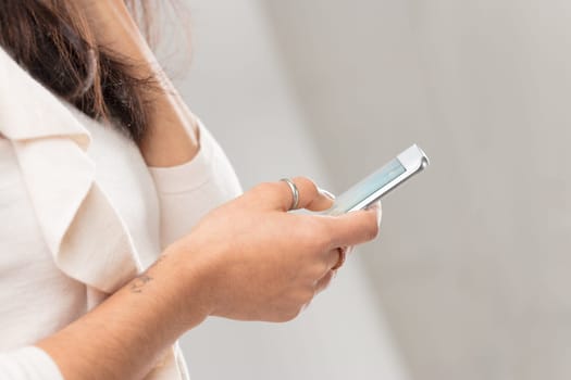 Close focus on the hand of a young woman clutching a smartphone on which she is scrolling. Image tones are warm and creamy white with lots of copyspace.