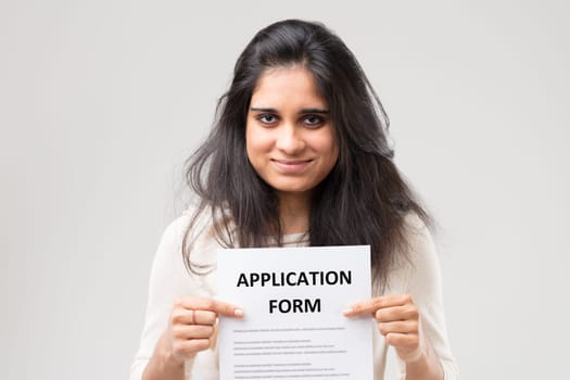 Horizontal half-length portrait of young Indian woman holding a sign with a paper application form in her hands. Her gaze seems to say, 'What are you waiting for to fill out the form? We are here!'