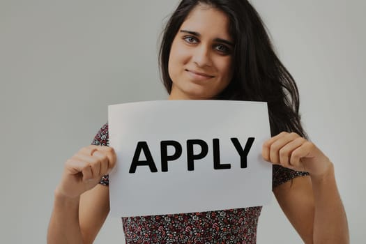 With a sign that simply reads 'APPLY,' a woman with striking eyes and a welcoming demeanor stands as the messenger. Her outfit's design accentuates the overall composition