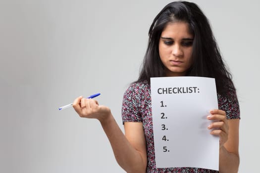 Young Indian woman or girl with long black hair and a flowery dress who could be as much a student as a novice worker. She holds a checklist in one hand, which she stares at analyzing it, and a pen in