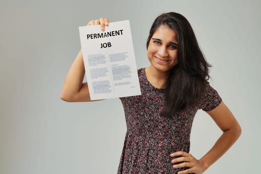 With a beaming smile and confident posture, a woman holds up a paper proclaiming 'PERMANENT JOB.' The floral pattern of her dress contrasts with the formality of the employment milestone