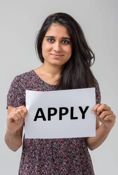 Portrait of an Indian girl in a flowery dress and long hair holding a sign. she is happy and smiling and the sign says 'apply,' inviting you because it is time for you to step up in the same company w