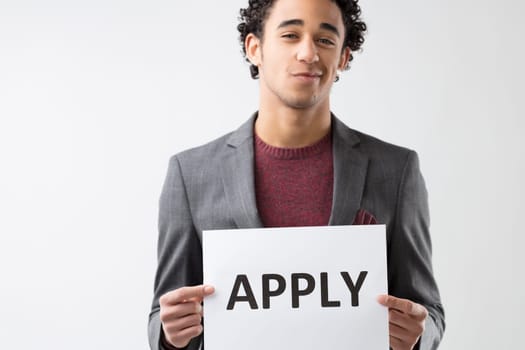 cheerful man with curly hair encourages with a sign reading 'APPLY,' dressed in a sophisticated gray suit and burgundy sweater