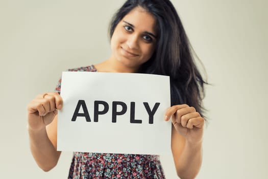 woman with expressive eyes and flowing dark hair confidently holds a sign stating 'APPLY.' The floral pattern of her attire contrasts the simple, clear message she's conveying