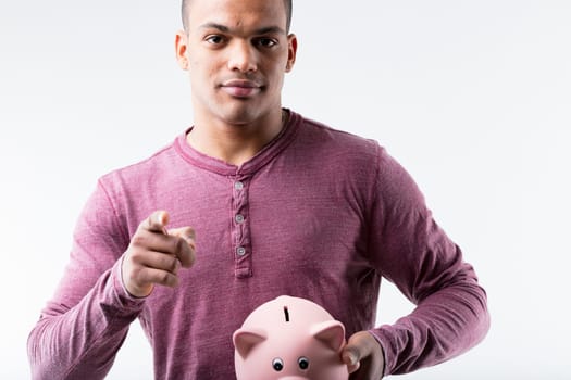 Black man, burgundy shirt, holding piggy bank, pointing at us. Encouraging to save for investing, or buying without wasting money. Set against a white background