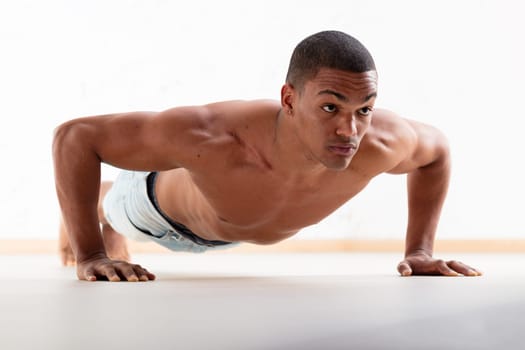 Black man does wide-arm push-ups. With his chest in shape and arms to the sides, this young man with short hair exercises shirtlessly on a light wooden floor, striving for great physical health