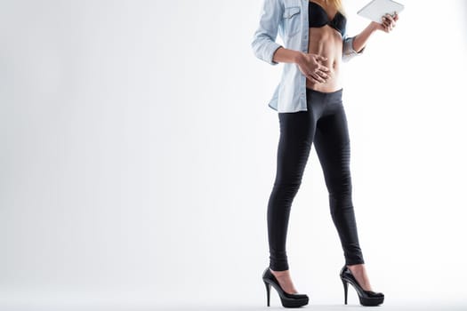 Fitness app concept: a fit unrecognizable woman with a toned body, flat stomach, in an open denim shirt, uses a digital tablet to manage workouts. White backdrop