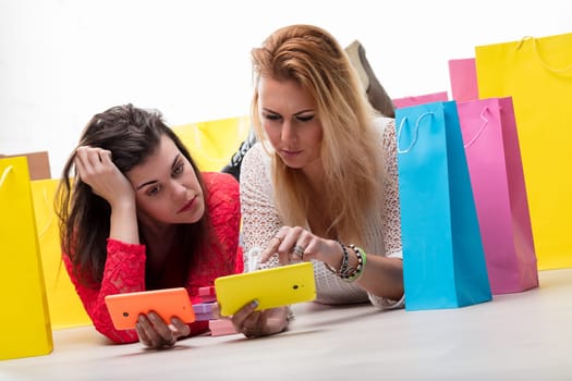 Two women enjoy a break after shopping, browsing on their phones amidst colorful bags