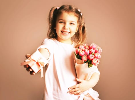 Portrait of a beautiful smiling little girl holding a bouquet of spring tulips and a gift box over a bright background. Happy Womens Day, Father's Day, Easter celebration.