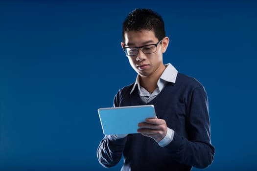 Asian man, glasses on, absorbed in his tablet, checks news against a blue backdrop. His interest lies in the screen's content - video, news, an email, or a security camera feed