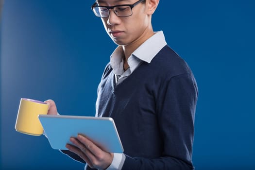 An Asian man checks news on his tablet, holding a yellow mug, against a blue backdrop. He's engrossed in the screen content, may it be a video, news, an email, or a security camera view