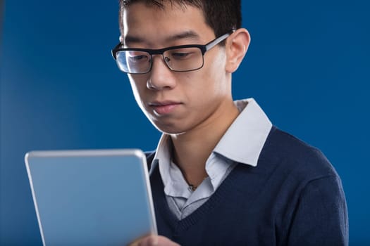 Asian man, absorbed, scrutinizes news on tablet, glasses on. Against a blue backdrop, he's engaged with the screen - video, news, email, or a security camera view