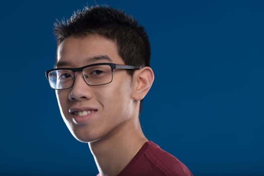 Portrait of a young East Asian man wearing glasses and smiling in a burgundy T-shirt. Short-haired, against a blue background, his image radiates future success and positivity