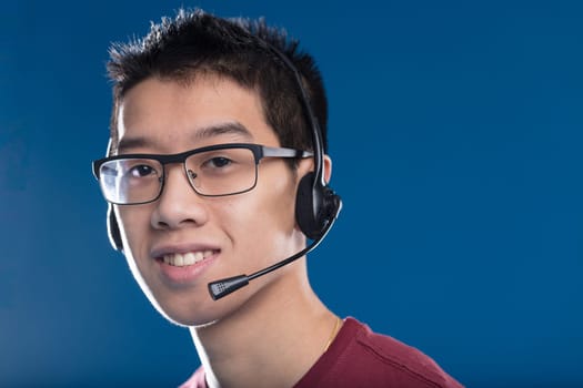 A young smiling Asian adult wears a typical gamer or call center headset and smiles helpfully. Gamers communicate with their team