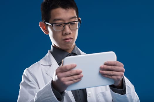 Asian scientist peruses updates and data on his tablet. A Chinese ethnicity researcher, set against a blue backdrop. Wearing glasses, intrigued by screen content - videos, news, emails, comparative st