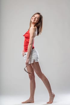 Confident young woman in red tank top, white shorts, holding sunglasses, slim and fit, not shy to show body at beach, neutral background