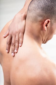 From the back, a fit man lifts his arm, hand nearing his neck's base. Are muscle strains, neck pain, or migraines a concern? His head is cleanly shaved his face remains unseen