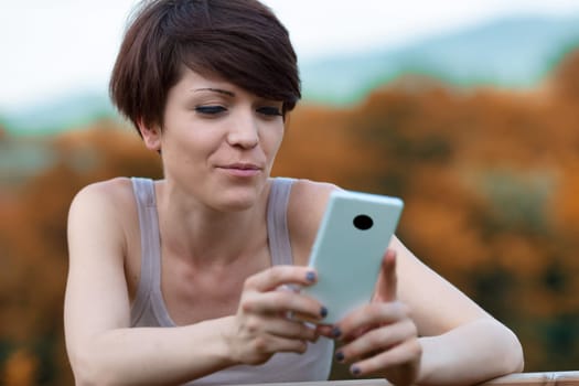 Young woman reading a text message on her mobile phone with a wry grin as she relaxes outdoors in autumn in a close up cropped view
