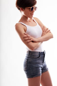 Sexy jaunty young woman in denim shorts and trendy sunglasses standing with folded arms and a quiet smile in a cropped side view isolated on white