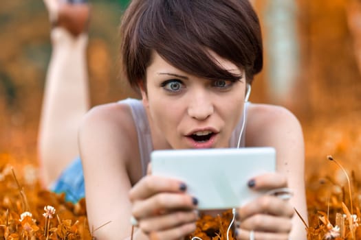 Young woman staring at her mobile in shock or amazement with wide staring eyes and open mouth as she lies outdoors in a meadow relaxing amongst wildflowers
