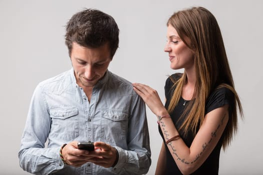 A man grins at his smartphone, girlfriend observing. Knowing his gaming habits, she accepts them lovingly. The relationship sustains despite shared flaws