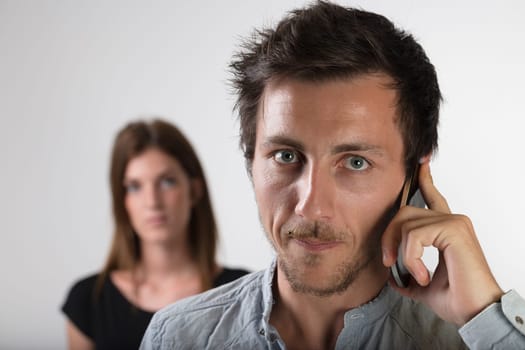 'In the foreground, a man is on a smartphone call a suspicious woman is blurred in the background. Betrayal, jealousy, love, tumultuous relationships. Concept of woman's jealousy and control over man