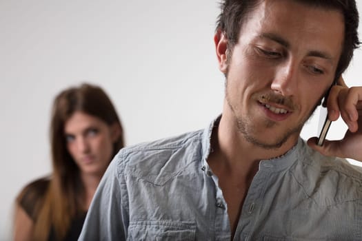 'In the foreground, a man is on a smartphone call a suspicious woman is blurred in the background. Betrayal, jealousy, love, tumultuous relationships. Concept of woman's jealousy and control over man