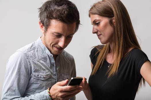 Man in light shirt smiles at smartphone his girlfriend watches lovingly. Aware he's viewing sexy content or gaming, she tolerates his habits out of love
