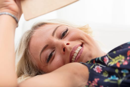 A young lady with blonde hair, a floral dress, pausing her book to grace us with a smile. Lying on a sofa, she looks beautifully made up