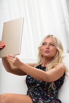In a white living room, a woman on a white sofa makes a duckface for a selfie on a large tablet, perfecting her pose. Vanity or social media slave?