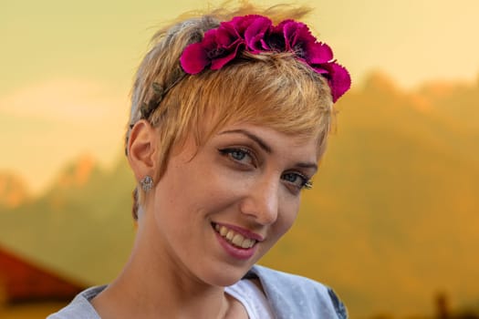 Close-up portrait of a woman with short blonde hair, tidy and made-up, wearing a headband made of fuchsia flowers, against an outdoor background that turns all orange, as she smiles happily in love wi