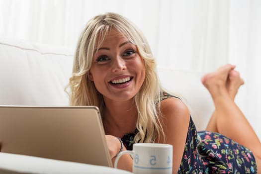 On her sofa, a happy blonde woman with a tablet smiles cheerfully. Could she be shopping, indulging in a TV show, or viewing a social media video