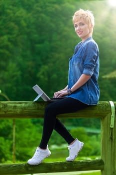 Stunning travel blogger and tourist sitting on a wooden fence in the middle of a mountain valley in Europe, writing and telling her followers about the fantastic adventure in tourism offered by the ol