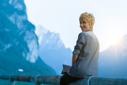 Stunning travel blogger and tourist sitting on a wooden fence in the middle of a mountain valley in Europe, writing and telling her followers about the fantastic adventure in tourism offered by the ol