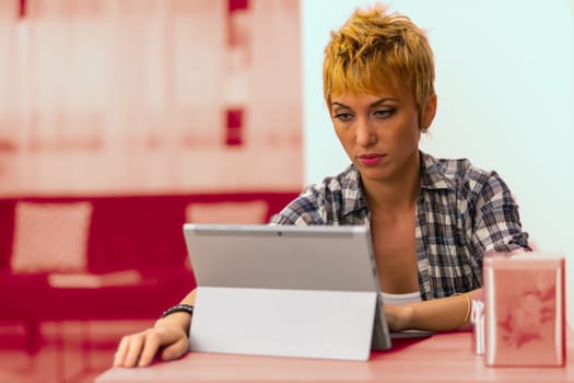 Freelance woman with short blonde hair and a plaid shirt works in a rustic public place with her laptop-tablet combo. Highly focused and concentrated on writing and researching online as a serious pro