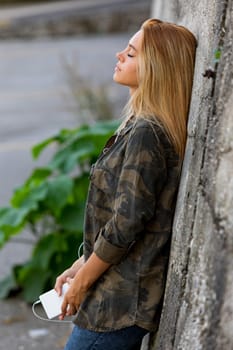 Gorgeous blonde girl leaning against a concrete wall in a small town, lost in thoughts while listening to something with wired earphones connected to her smartphone, like music or a podcast. She wears