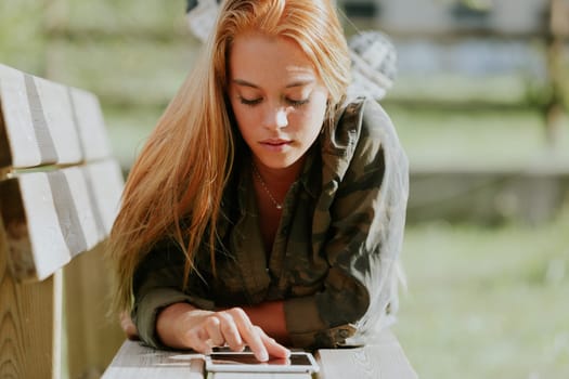 Beautiful blonde girl in casual-sporty attire fiddles with a digital tablet while lying on a park bench, using the internet, studying, working, or having fun