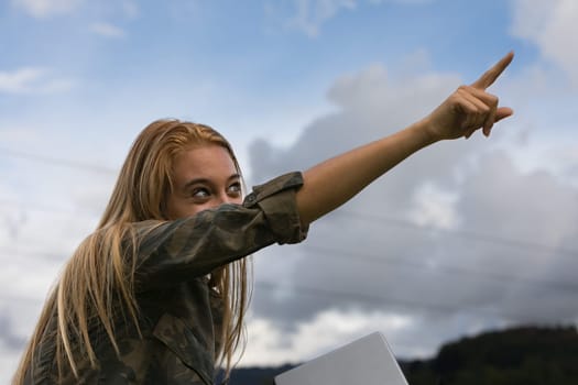 gaze of an androgynous girl in a young blond woman points to the sky with her index finger, but behind her are high voltage cables carrying electricity she has a tablet in her hand