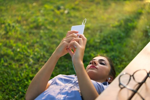 A three-quarter view from above shows a girl lying on her back on a bench, with wired earphones connected to her smartphone as she concentrates on the screen. She may be engaged in a video call or enj
