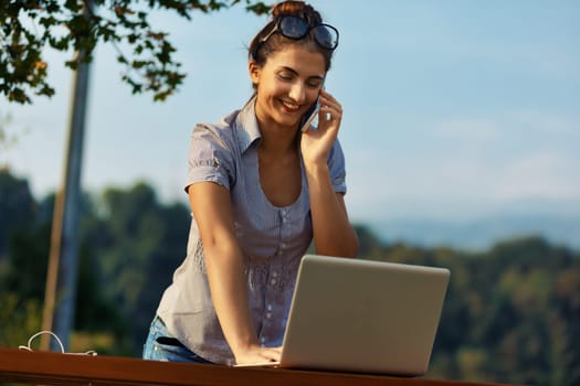 Young lady working, talking animatedly outdoors. Juggling phone and laptop, adorned in striped blouse. Glasses keep her hair, backdrop of towering mountains. She smiles, engaged in lively conversation