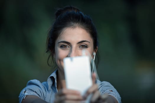 A young woman with brown hair takes selfies or makes a video call using her white smartphone. She extends her arms to improve the framing while wearing wired earphones. She is in a public park surroun